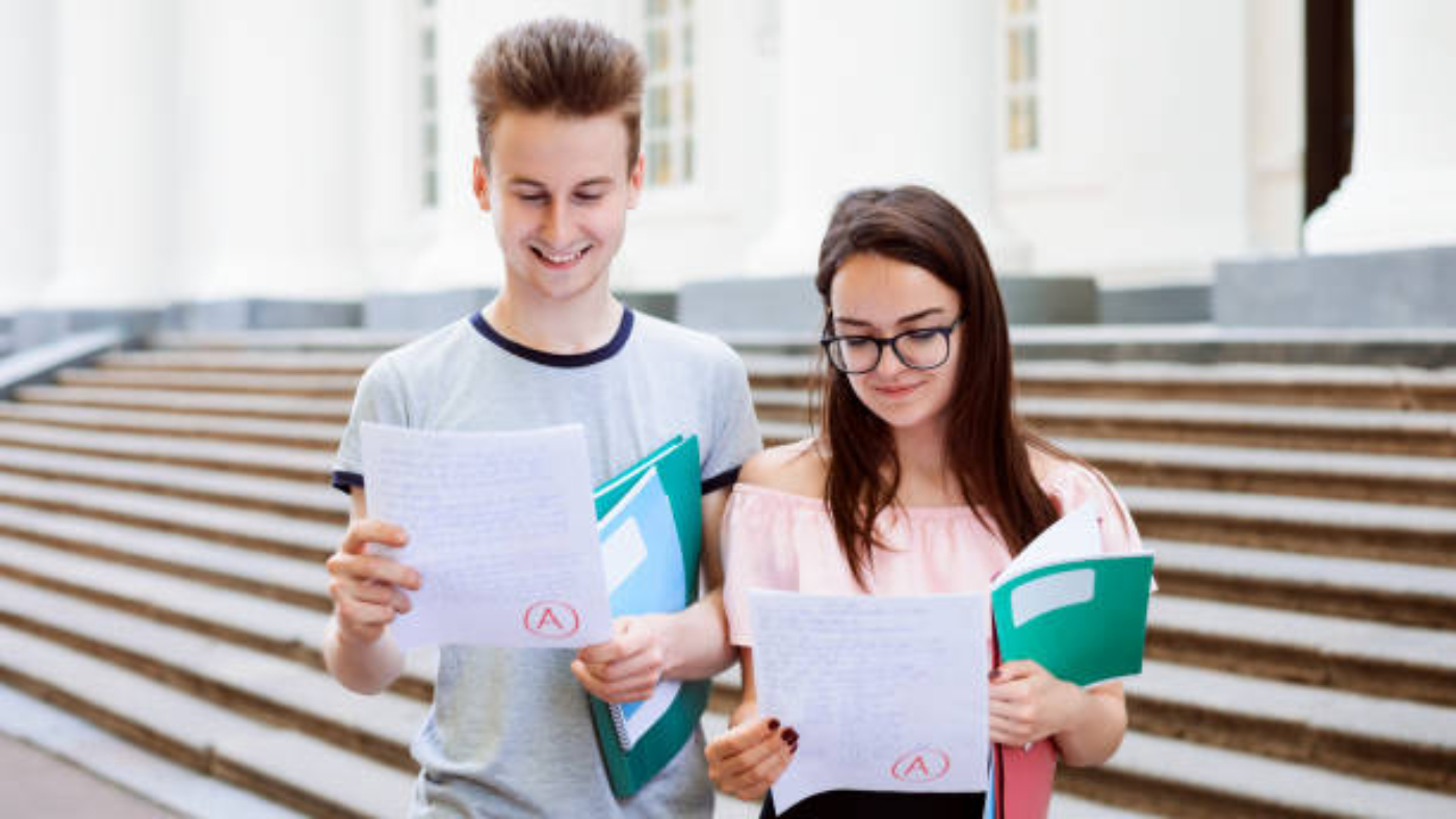 Male and female teenagers standing in front of university looking at excellent results of exam, happy to get the best mark, holding folders and books