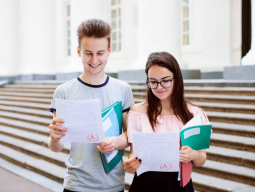 Male and female teenagers standing in front of university looking at excellent results of exam, happy to get the best mark, holding folders and books