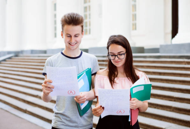 Male and female teenagers standing in front of university looking at excellent results of exam, happy to get the best mark, holding folders and books