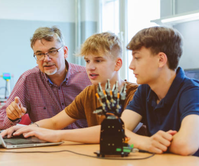 Gen Z teenage boys high school students with their teacher, coding movement of robotic hand at high school computer lab.